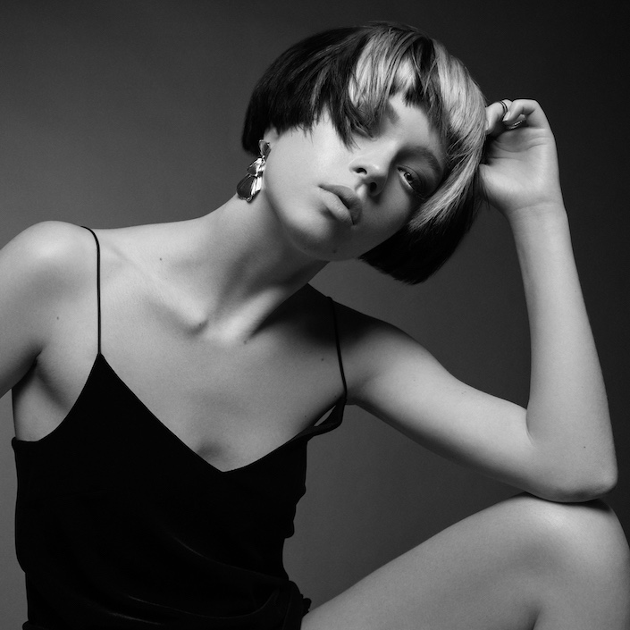 A black an white I mage of a female model with a short dark bob and blonde fringe sitting down resting her elbow on her knee